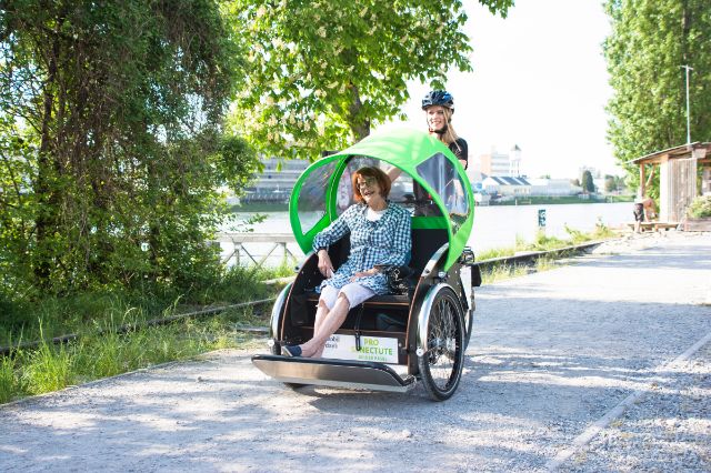 Seniorin und junge Frau auf einem Ausflug entlang des Rheins mit einer E-Rikscha.